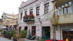 Un edificio histórico con detalles arquitectónicos ornamentales, un balcón con plantas en macetas y asientos exteriores con cojines rojos.