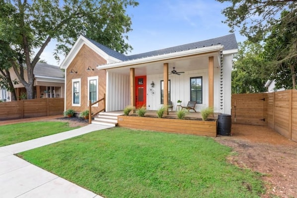 A single-story house with a red front door, a covered porch with a ceiling fan, and a small garden area.