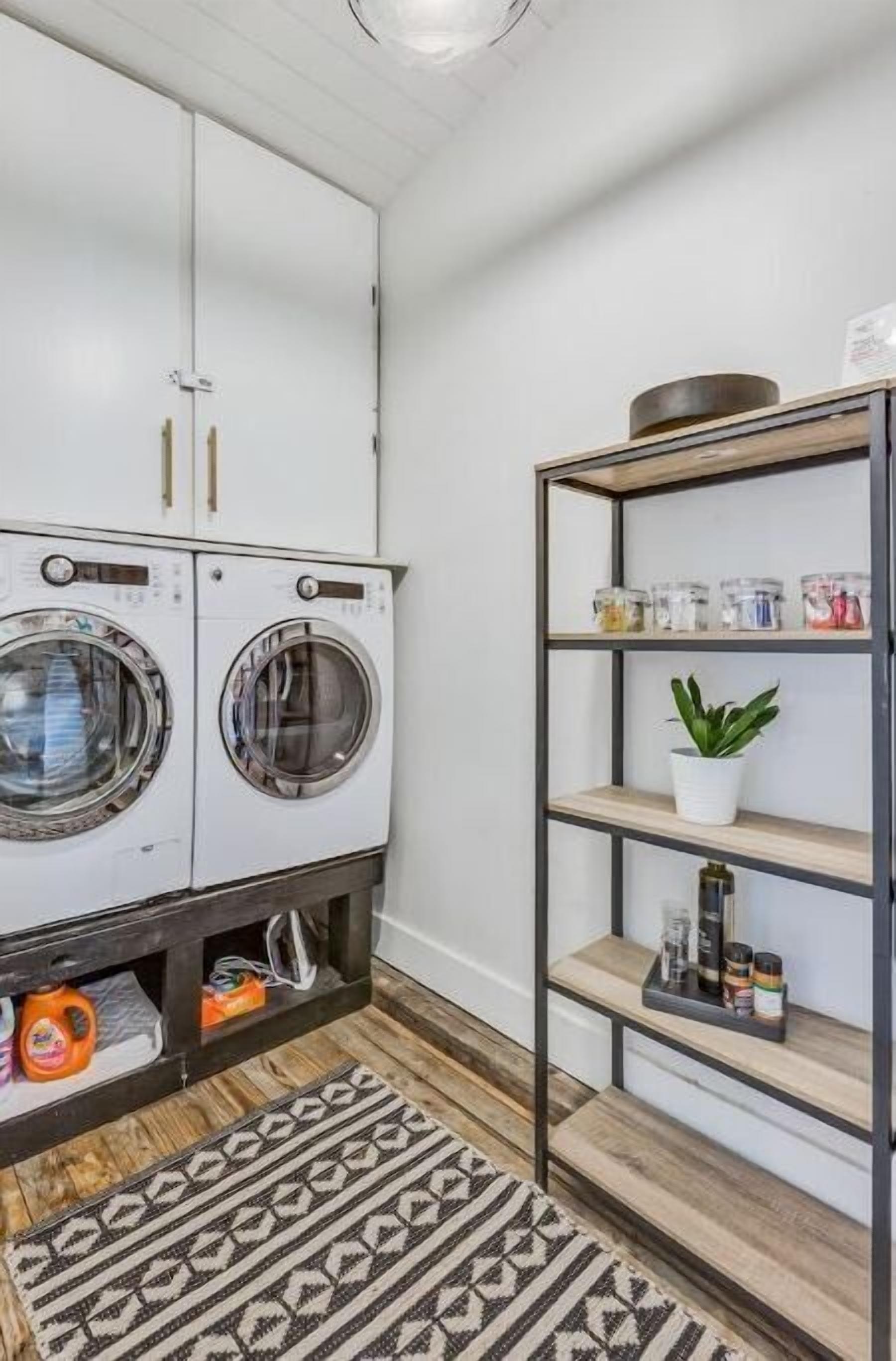 A laundry room with two front-loading washing machines, a shelf with storage containers, and a potted plant.