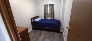 A bedroom with a single bed, a wooden headboard, a window with curtains, and a door.