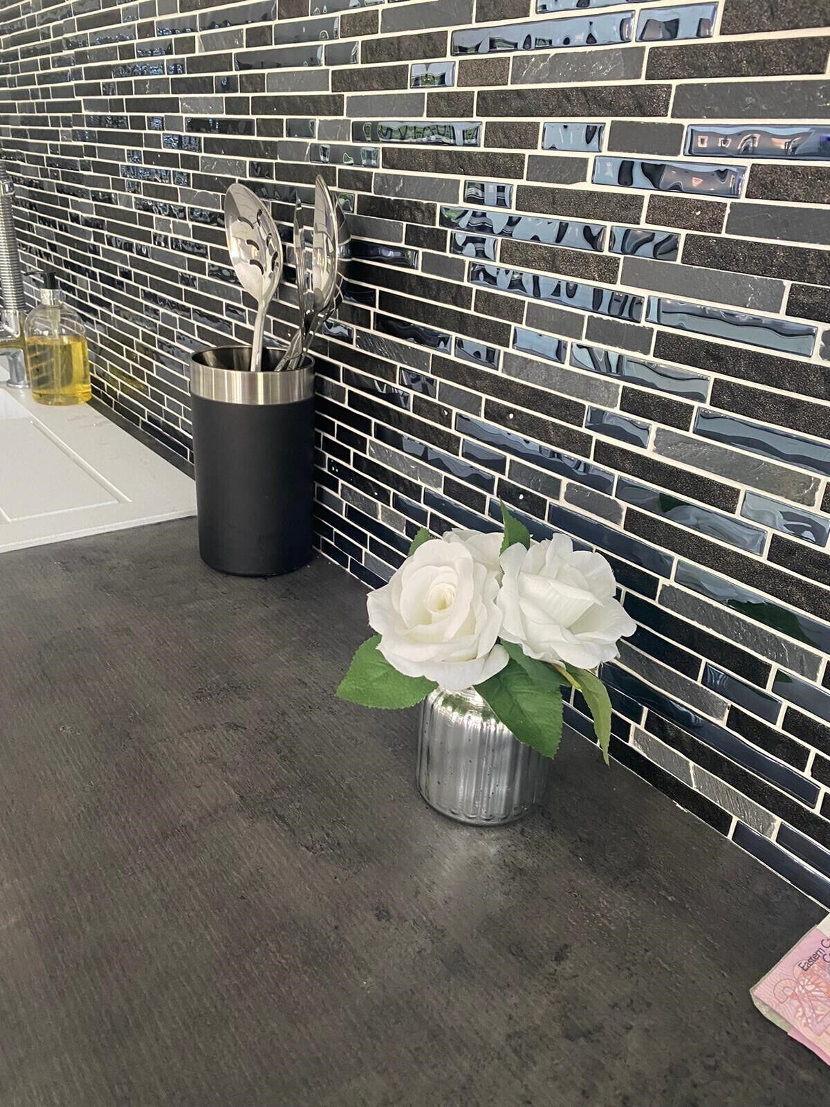 Ein Badezimmer mit schwarz-weißer Fliesenrückwand, einem Edelstahlschalenhalter und einer Vase mit weißen Blumen auf der Arbeitsfläche.