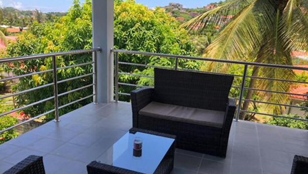 A balcony with wicker furniture, a glass table, and a view of lush greenery and distant buildings. - Stunning 3-bed Apartment in Rodney Bay (Gros Islet)