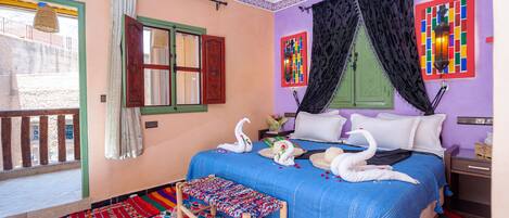 A room with a bed, a bench, a window with curtains, a wall with colorful artwork, and a balcony.