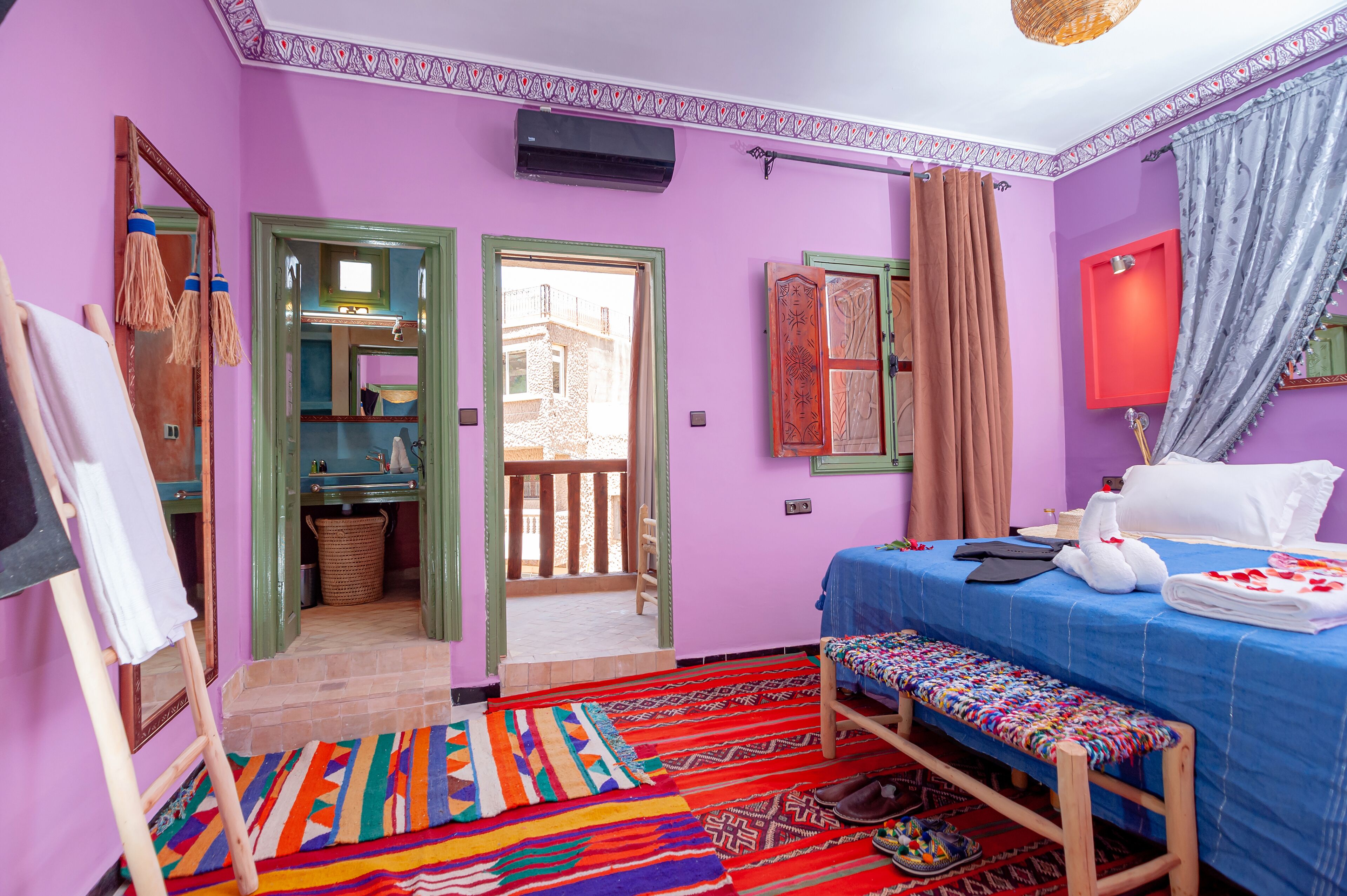 A room with a bed, a bench, a colorful rug, a mirror, and a view of the outside.