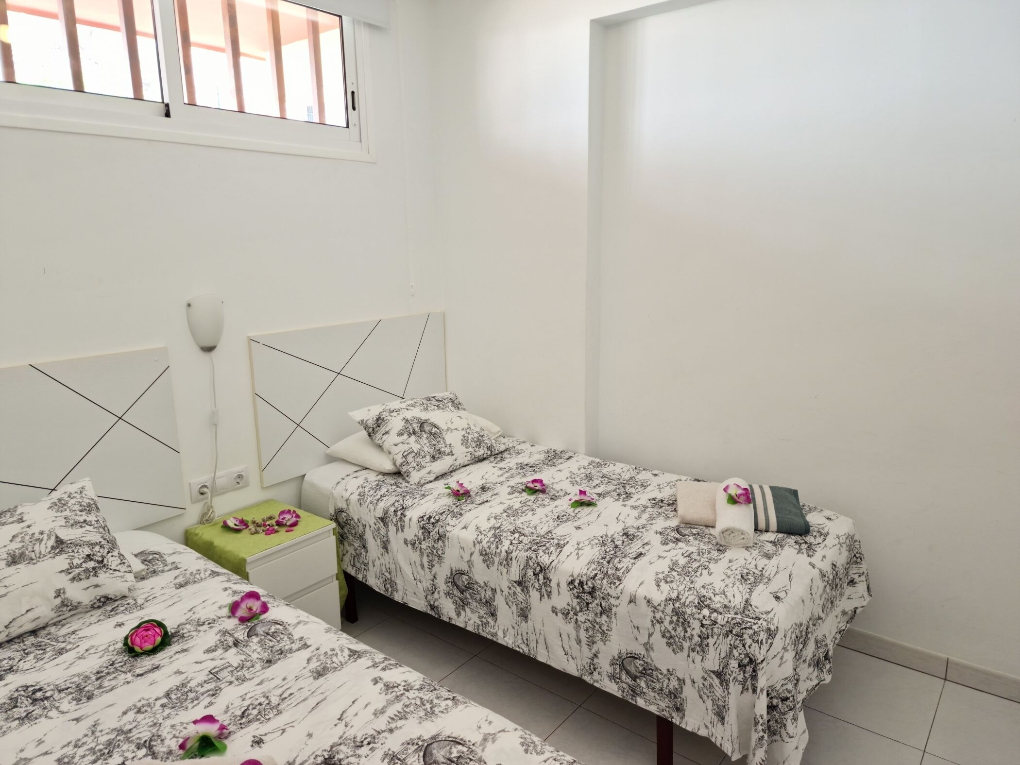 A bedroom with two beds, a bedside table, a nightstand, and a window.