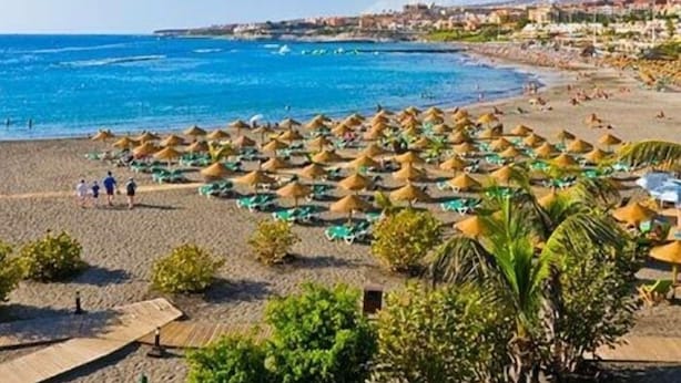 Ein Strand mit Reihen von Sonnenschirmen und Liegestühlen, einem klaren blauen Meer und Gebäuden im Hintergrund.