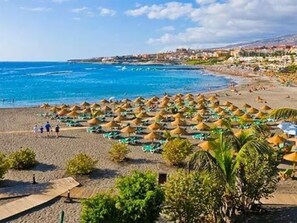 Una playa con hileras de sombrillas y tumbonas, un mar azul cristalino y edificios al fondo.
