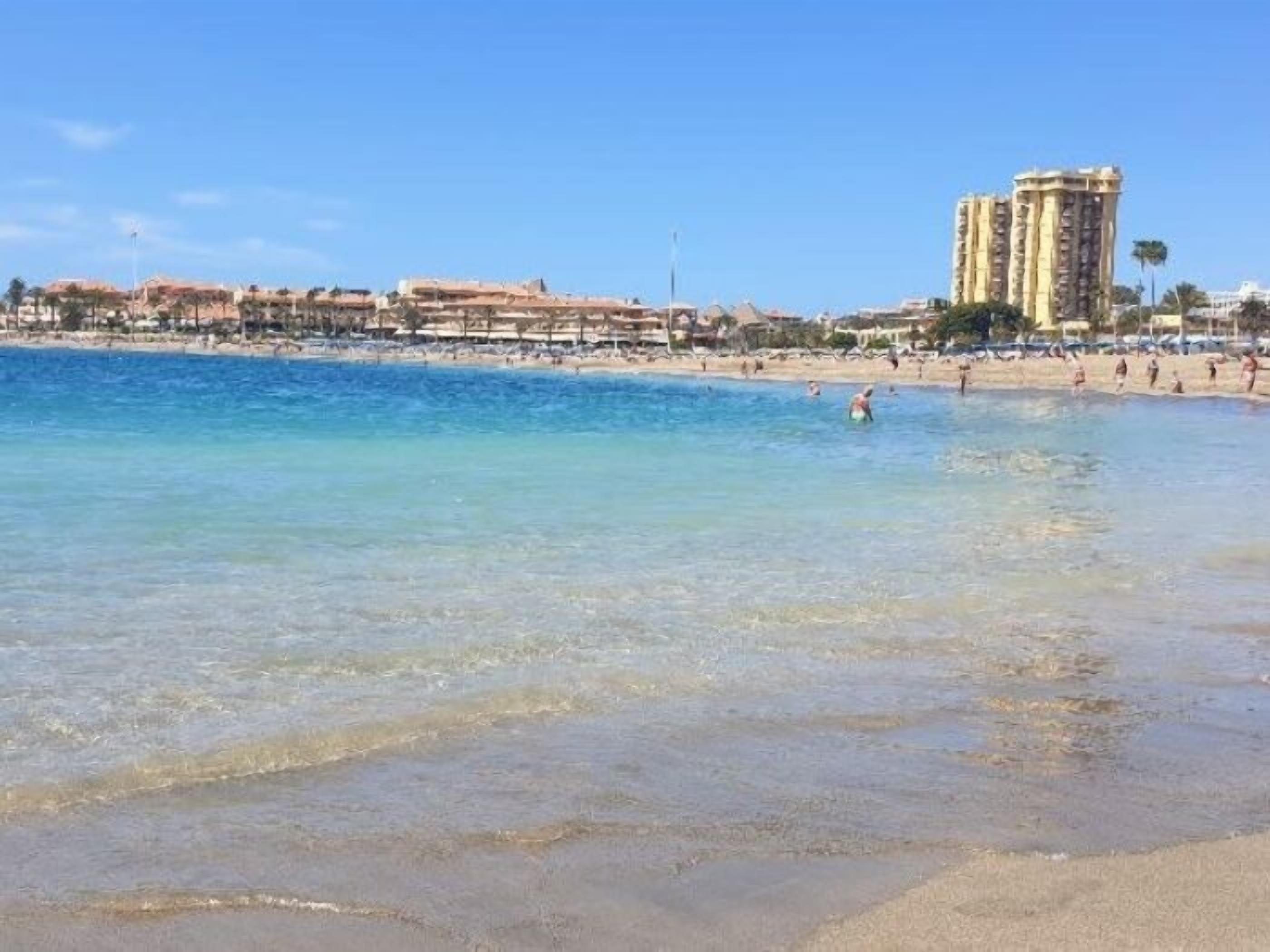 A beach with clear blue water, a sandy shore, and a tall hotel building in the background.