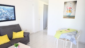 Ein Wohnzimmer mit grauem Sofa, gelben Kissen, einem weißen Couchtisch mit Pflanze, einem Esstisch mit grüner Tischdecke und weißen Stühlen sowie einem Stadtbild an der Wand.