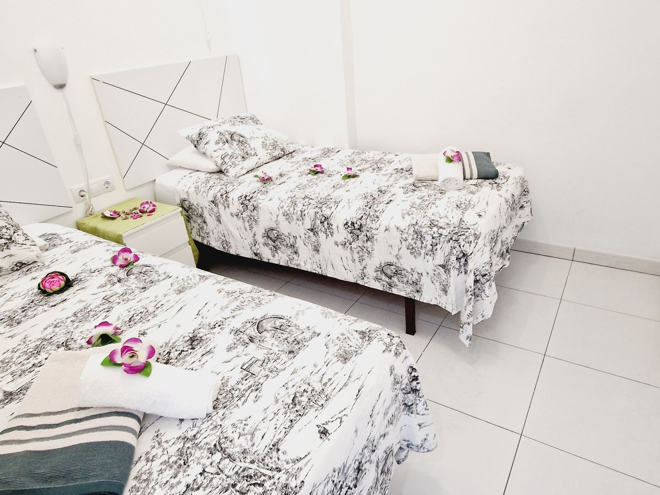 Una camera d'albergo con due letti, un comodino con lampada e un tavolino con un vaso di fiori.