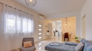 Un salon moderne avec un canapé gris, une chaise en bois et une table basse blanche. On y trouve également un coin repas avec une table et des chaises, et une cuisine en arrière-plan.