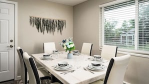 Comedor con una mesa dispuesta para seis comensales, sillas blancas, un jarrón con flores y un adorno en la pared.