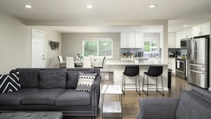 Una sala moderna con un sofá gris, una mesa de centro y una zona de comedor con una mesa y sillas blancas. Se ve la cocina, que cuenta con electrodomésticos de acero inoxidable y armarios blancos.