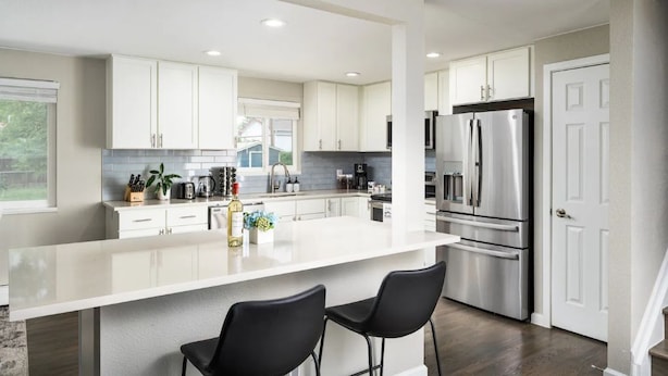 Una cocina moderna con armarios blancos, electrodomésticos de acero inoxidable, una isla grande con dos taburetes negros y vistas al exterior a través de un ventanal.