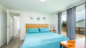 A bedroom with a wooden headboard, a bed with a blue patterned bedspread, a nightstand with a lamp, and a view of the outdoors through large windows.