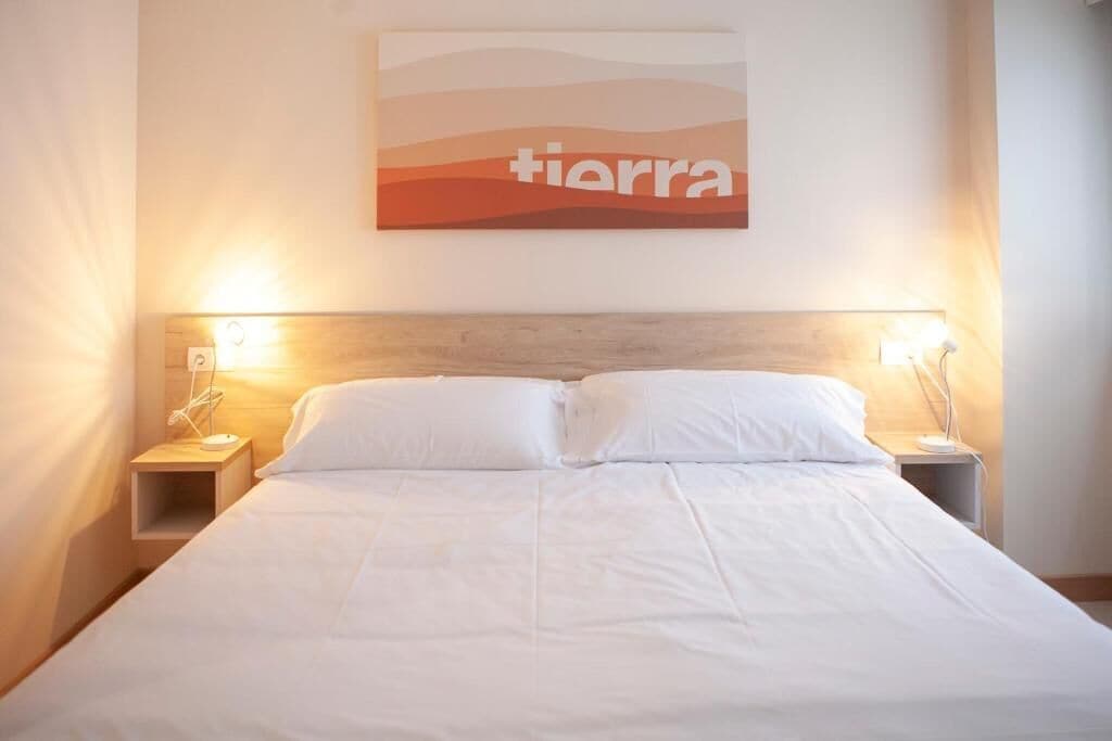 A neatly made bed with white linens, two bedside tables with lamps, and a wall-mounted artwork with the word "tiarra".