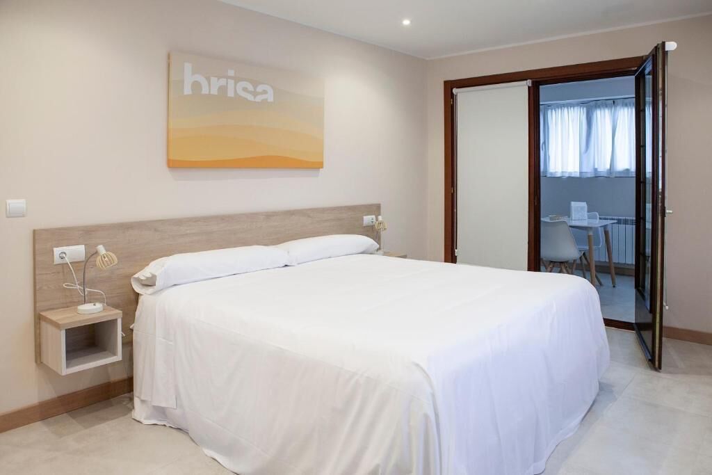 Habitación de hotel con una cama grande, una mesita de noche con lámpara, un estante pequeño y una puerta corredera que da a un balcón con una silla y una mesa.