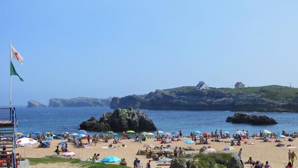 A beach with people sunbathing and swimming, a flag, and rocky formations in the background.