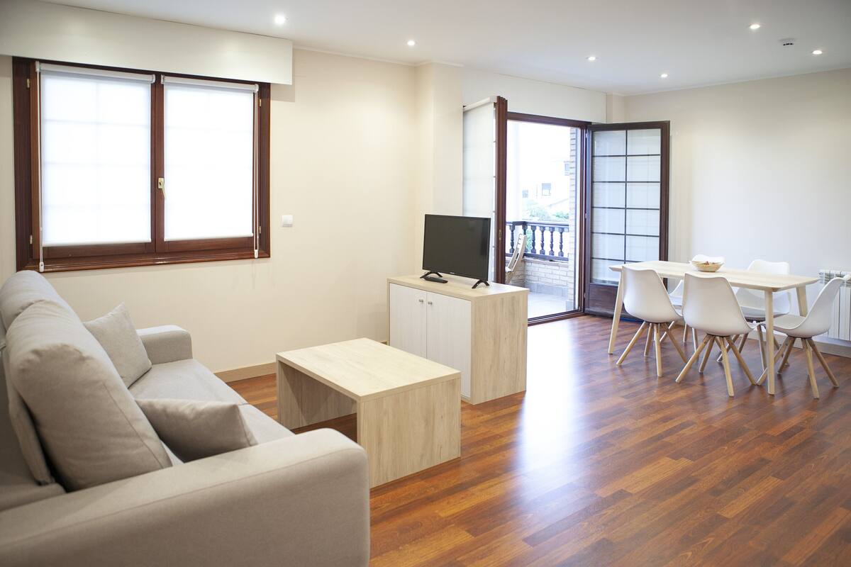 Un salon moderne avec un canapé, un téléviseur posé sur un meuble, une table à manger avec des chaises et un balcon doté d’une balustrade.