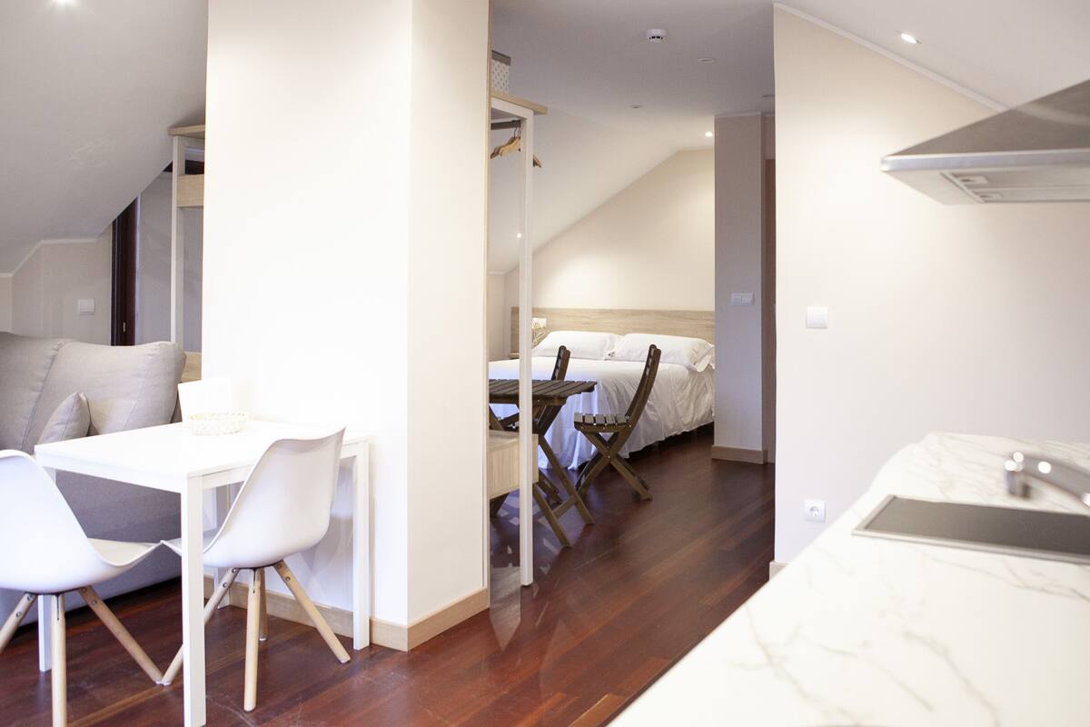 Une chambre moderne avec un plancher en bois, des murs blancs, un lit avec une literie blanche, une petite table à manger avec des chaises blanches et un comptoir en marbre blanc.