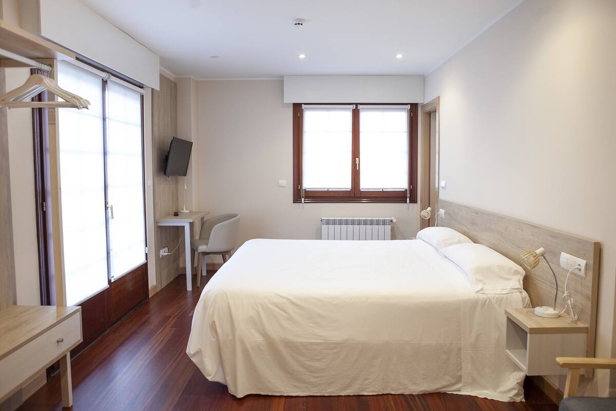 Une chambre à coucher comprenant un lit, une table de chevet, une chaise, un bureau, une télévision et une fenêtre avec des stores.