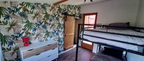 Une chambre avec un lit superposé, une commode blanche à tiroirs, une lampe rouge et une porte avec un rideau rouge.