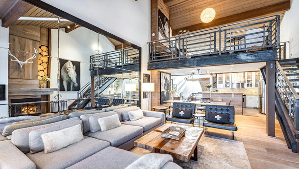 A spacious living area with a large sectional sofa, a wooden coffee table, and a fireplace. There is a staircase leading to an upper level with a balcony.