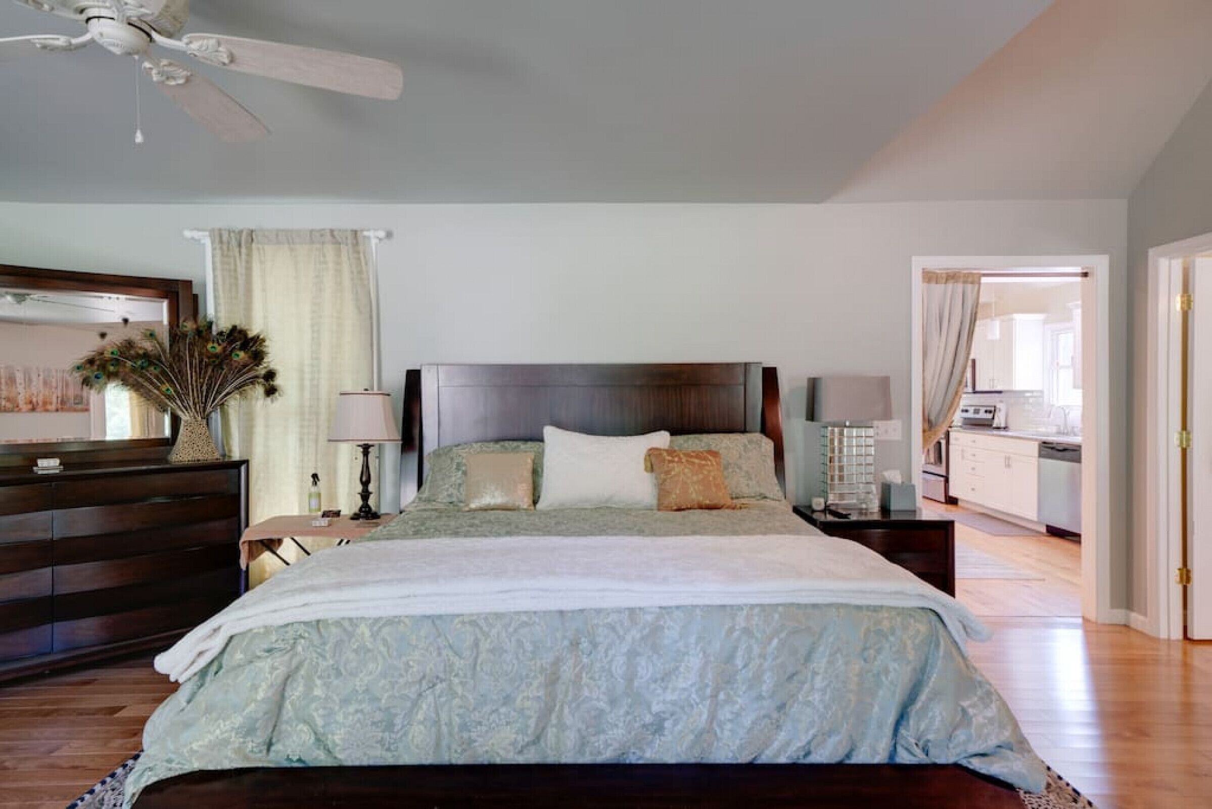 Une chambre à coucher avec un grand lit, une commode, une table de chevet, un ventilateur de plafond et une fenêtre avec des rideaux.