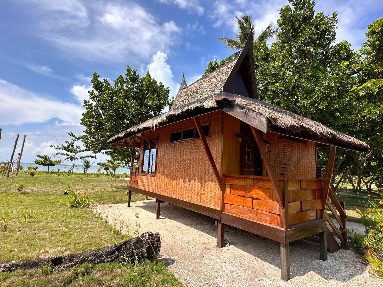 A thatched-roof hut with a wooden exterior, situated on a gravel path with a grassy area and trees in the background.