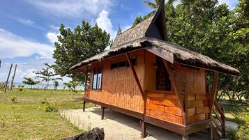 A thatched-roof hut with a wooden exterior, situated on a gravel path with a grassy area and trees in the background.