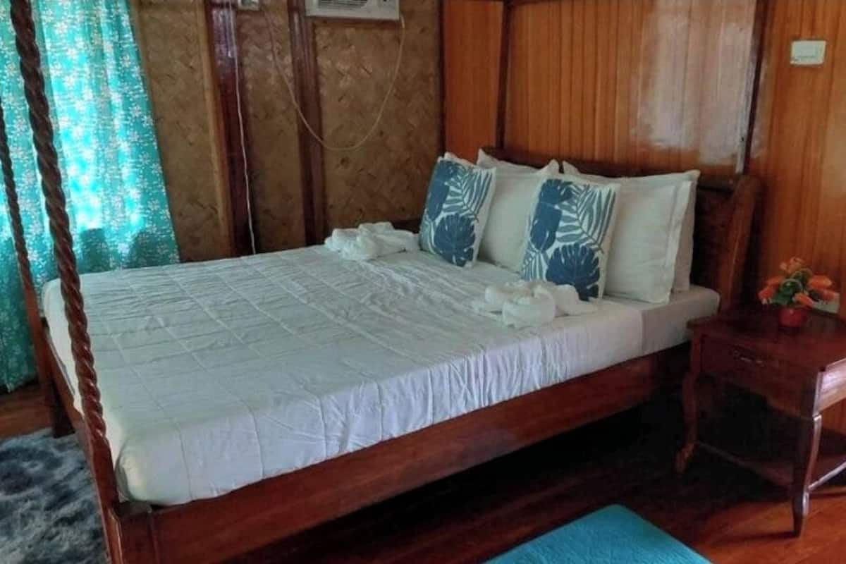 Een houten bed met wit beddengoed en blauw gestreepte kussens, een klein houten nachtkastje met een bloevas, en een blauwe mat op de vloer.