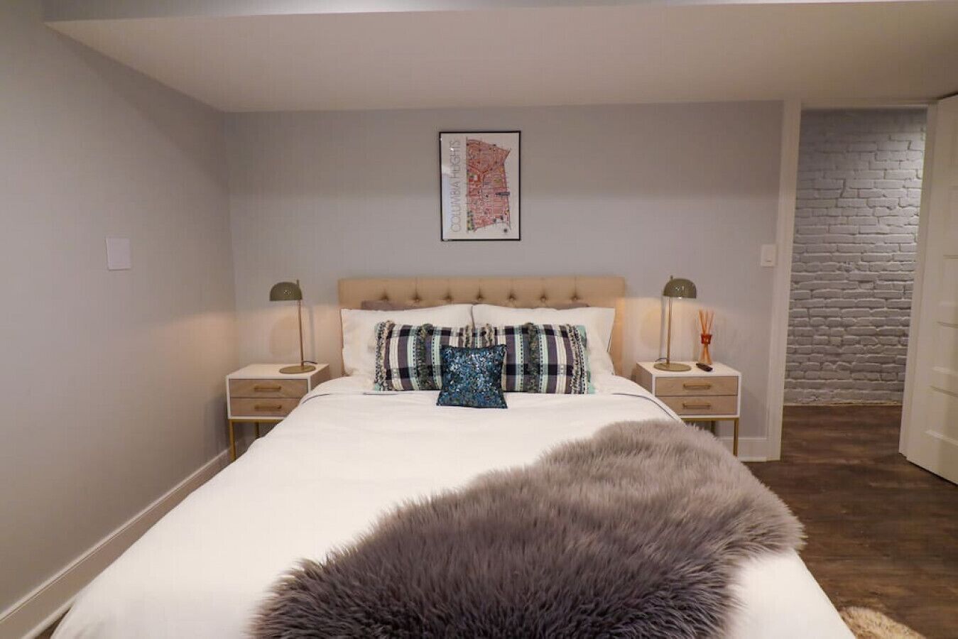 A bedroom with a large bed, two bedside tables, a framed map on the wall, and a brick wall in the background.