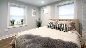 Een slaapkamer met een groot bed, twee ramen met jaloezieën, een deur en een klein plantje op de vensterbank.