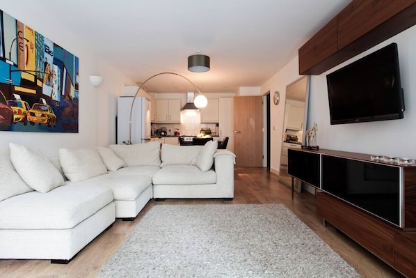 Ein modernes Wohnzimmer mit einem Ecksofa, einem Flachbildfernseher und einer Küche im Hintergrund.