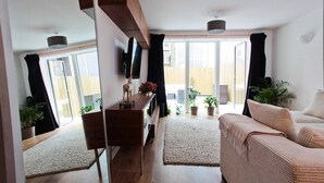 Ein modernes Wohnzimmer mit einer Couch, einem Couchtisch und einem Fernseher. Übertöpfe mit Pflanzen und eine Glastür, die zu einem Außenbereich führt.