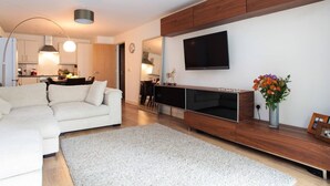 Ein modernes Wohnzimmer mit einer Couch, einem Fernseher, einer Holzunterhaltungsecke und einer Küche im Hintergrund.