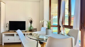 A modern living room with a glass dining table, white chairs, a flat-screen TV, a small potted plant, and a view of the outdoors through large windows.