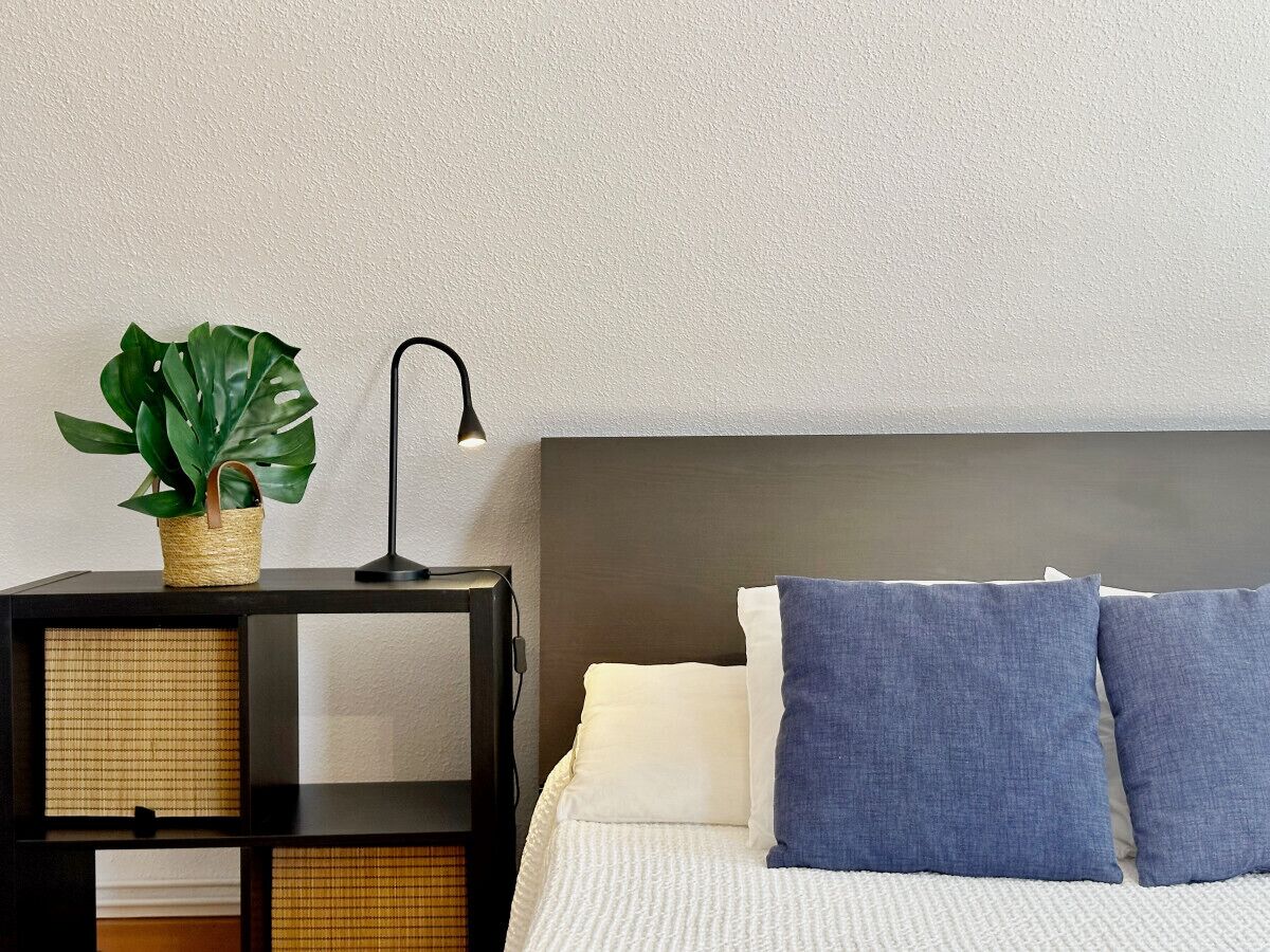 A neatly arranged bedroom with a bed, pillows, a nightstand, a lamp, and a plant.