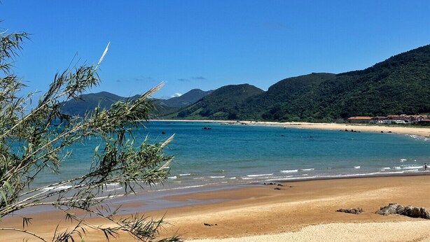 A beach with clear blue water, sandy shore, and green hills in the background.