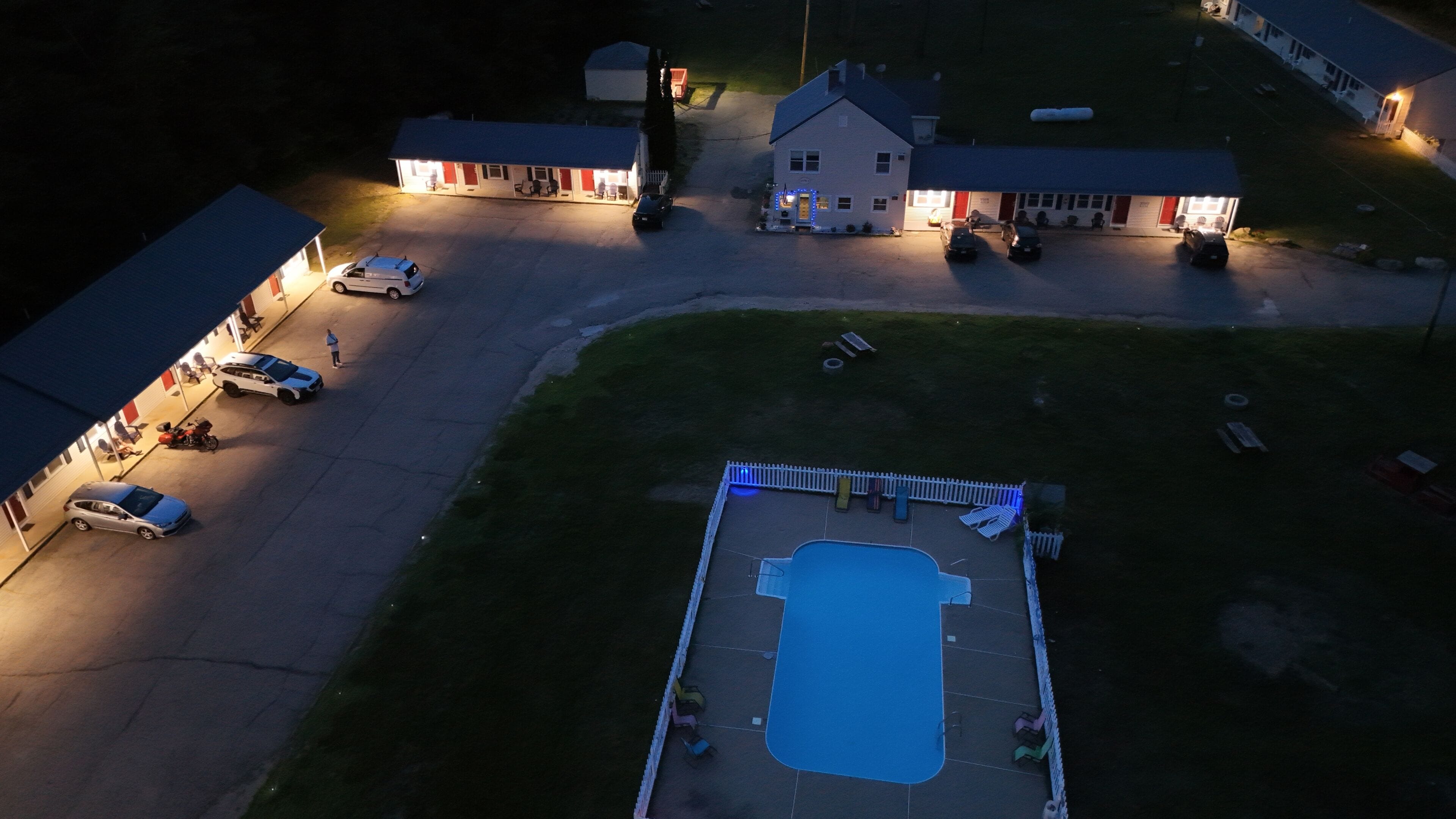 An aerial view of a property with a swimming pool, multiple buildings, and parked cars.