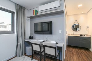 A modern room with a desk, two chairs, a flat-screen TV, a small kitchen area, and a large window with curtains.