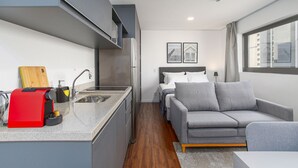 Un espace de vie compact comprenant un coin cuisine, un canapé gris et un lit avec une tête de lit.