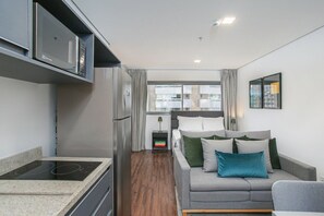 A modern kitchen with a built-in oven and microwave, a grey sofa with green and blue cushions, and a small bedside table with a lamp. - Arte Arquitetura Vila Mariana (São Paulo)