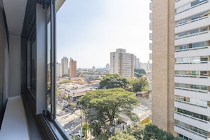 Um edifício alto com vários andares e varandas, uma grande árvore em primeiro plano e paisagem urbana ao fundo.