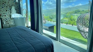A bedroom with a large bed, a bathtub, and a view of a green landscape through large windows. - Luxury Tiny home with pool & amazing view (Caguas)