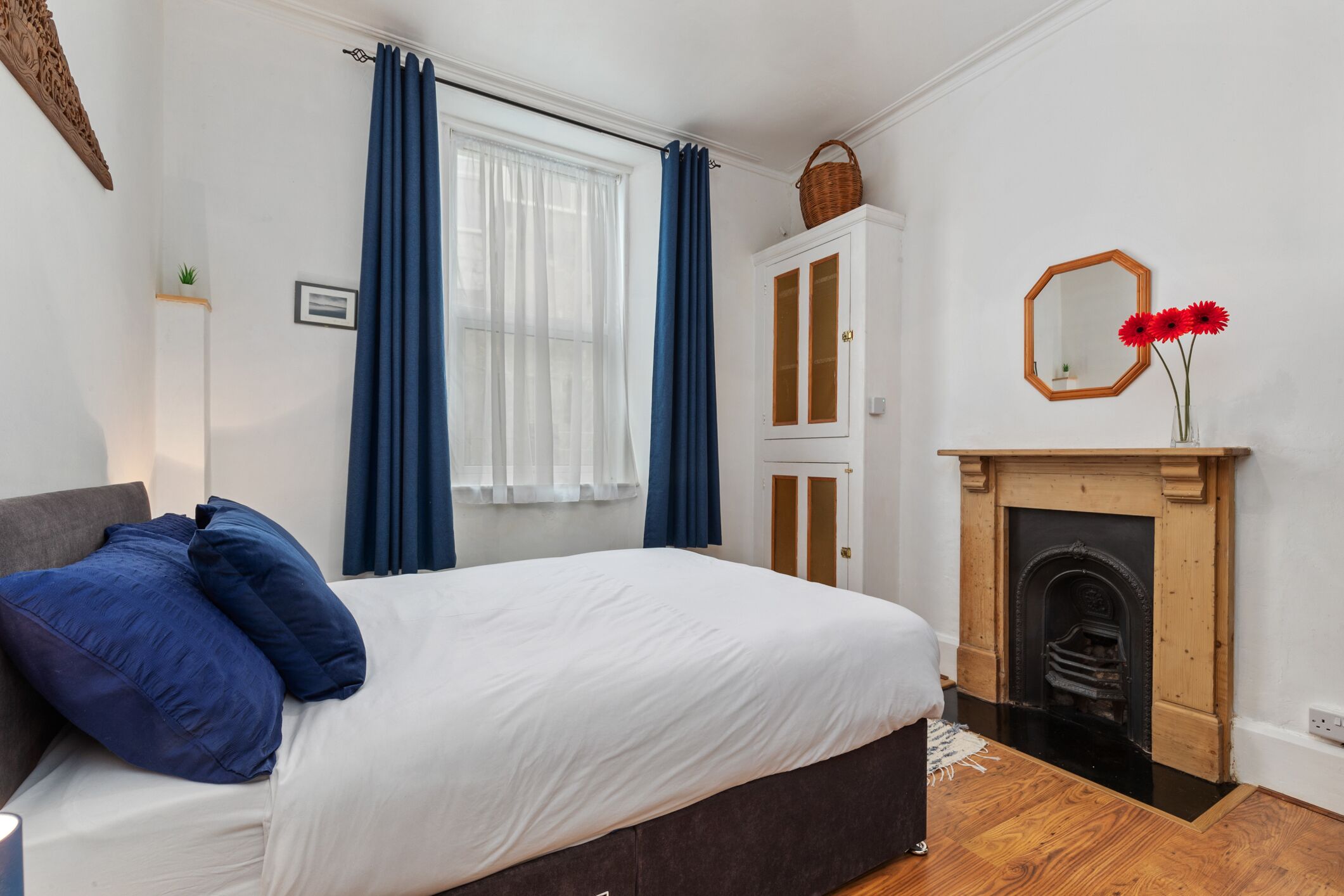 A bedroom with a bed, blue curtains, a wooden cabinet, a fireplace, and a mirror on the wall.