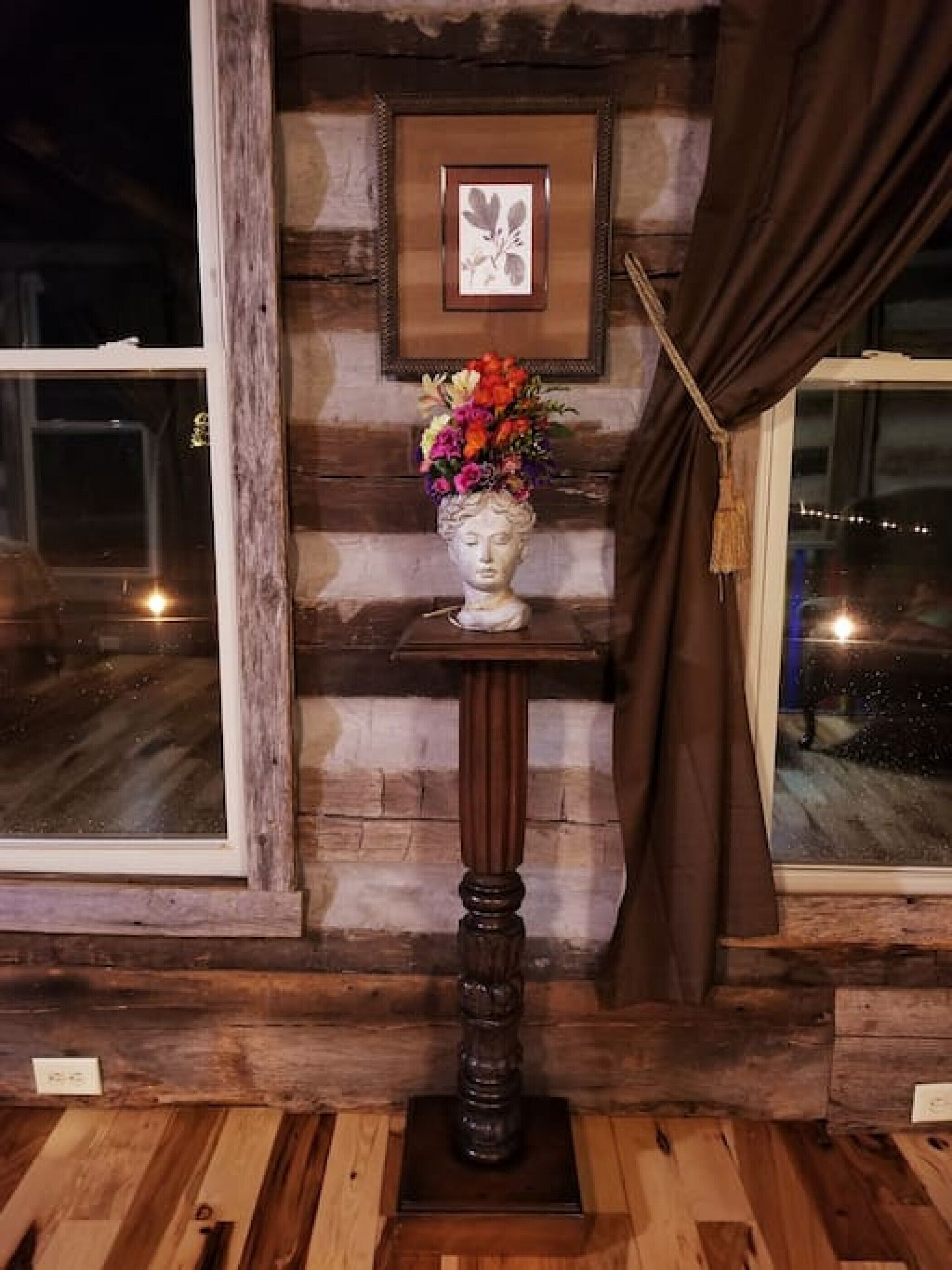 Un interior rústico con suelo de madera, pared de piedra, un cuadro enmarcado y un jarrón con flores sobre un pedestal.