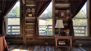 Interior rústico de una cabaña con paredes y piso de madera, y ventanas grandes con cortinas. Hay una pequeña mesa con una lámpara y un jarrón de flores.