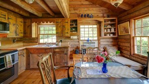Ein rustikales Blockhaus im Inneren mit Holzwänden, einer Essecke mit Tisch und Stühlen, einer Küche mit Holzausstattung und einem Spülbecken sowie einem Fenster, das für natürliches Licht sorgt.