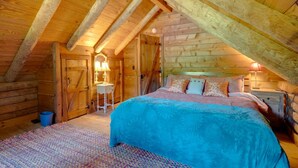 Ein gemütliches Schlafzimmer im Dachgeschoss mit Holzbalkendecke, einem großen Bett und einem kleinen Schreibtisch mit Lampe.